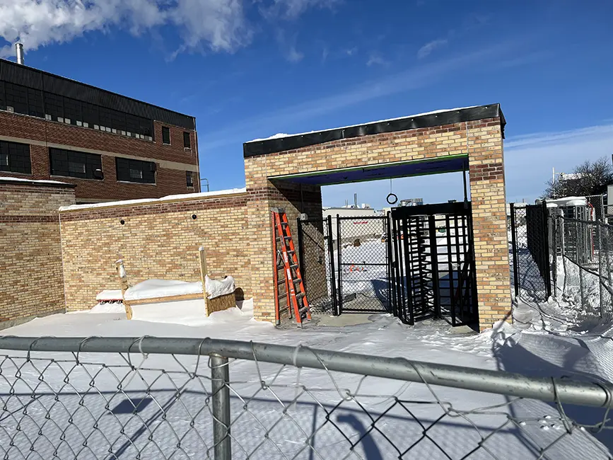 Outdoor construction area with a brick structure featuring a turnstile gate and a covered entryway. A red ladder leans against the brick wall, and snow covers the ground and surrounding surfaces. A chain-link fence encloses the area, with a larger brick building visible in the background under a clear blue sky.