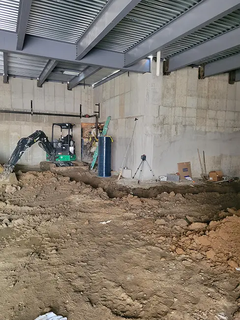 Interior construction site showing a mini excavator with a green body working in a partially enclosed space. The area has concrete block walls, metal ceiling beams, and exposed dirt floor with debris. Construction equipment and supplies are visible along the walls.