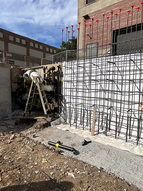 A construction site showing reinforcement steel bars (rebar) being installed against a brick wall. Red pole markers line the top of the wall. Construction equipment including a ladder and piping system is visible in the work area. The ground is dirt and concrete, indicating ongoing construction work.
