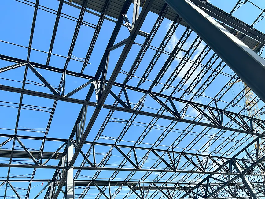 Steel truss framework of a building under construction against a bright blue sky.