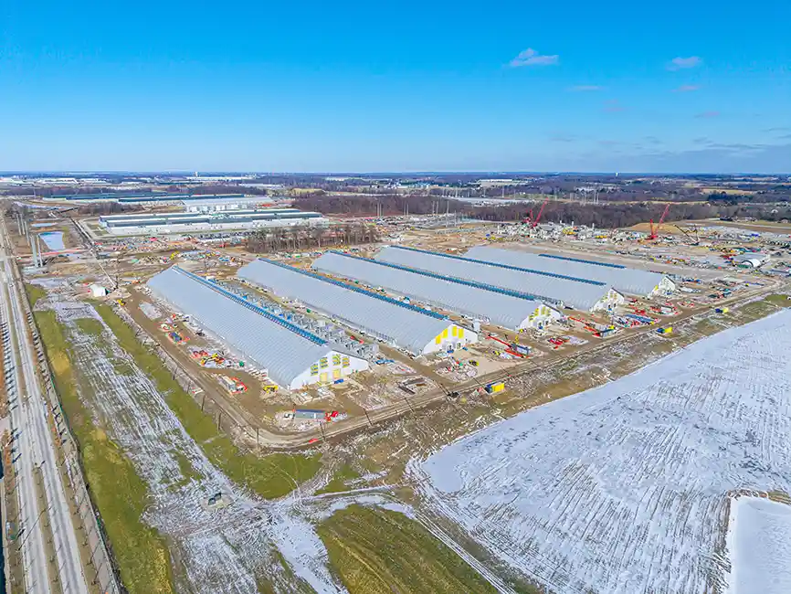Aerial view of a large construction site with multiple long, partially enclosed structures and surrounding equipment on a snowy landscape.