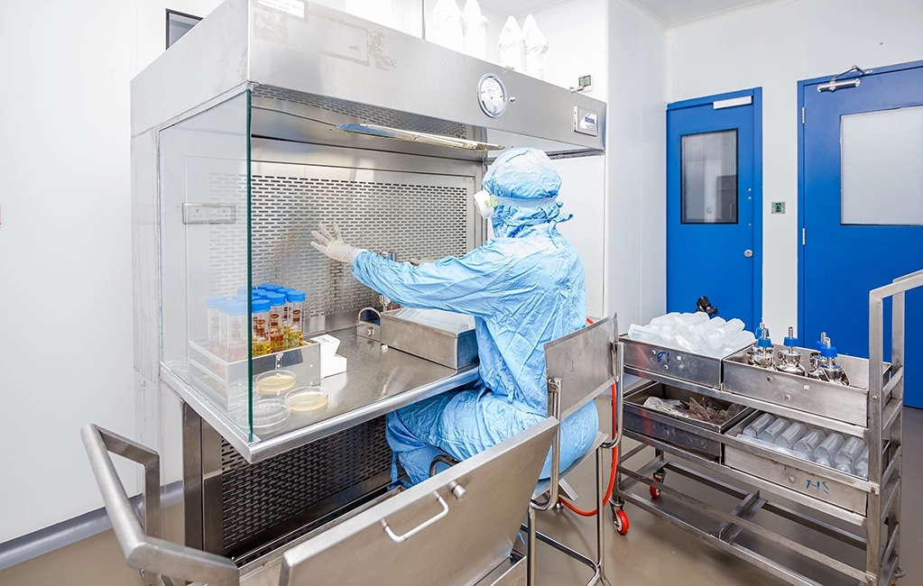 Person wearing sterile cleanroom garments seated at a laminar flow hood while preparing materials in a controlled laboratory environment.