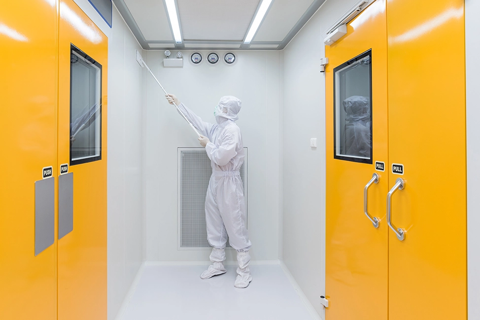 Person in full cleanroom garments cleaning the ceiling area of a controlled room with bright orange doors.