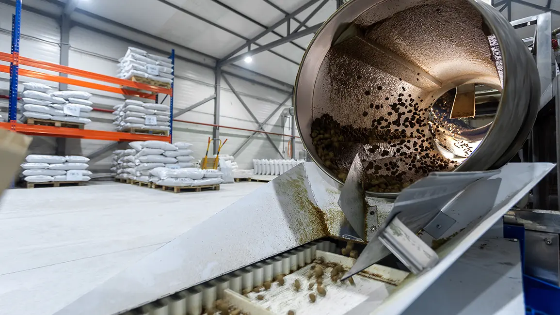 Large rotating drum tumbler coats pet food on a conveyor line inside a modern manufacturing facility, with ingredient bags stored on warehouse racking in the background.